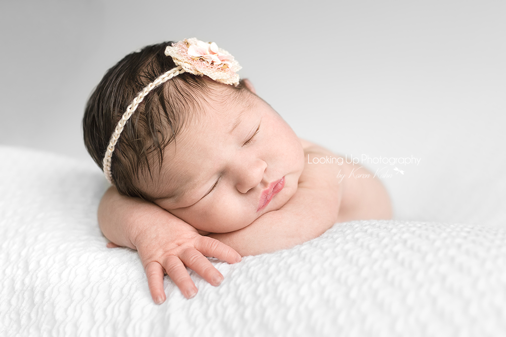 Sleepy baby girl posed reclining on arms with lovely hair and flower headband and pouty lips on white blanket for newborn portrait session