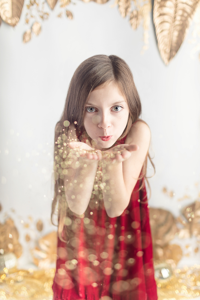 Girl with long hair and big eyes and festive red dress blowing glitter in the air in whimsical Joyful Gatherings gold set design for fall holiday portrait session