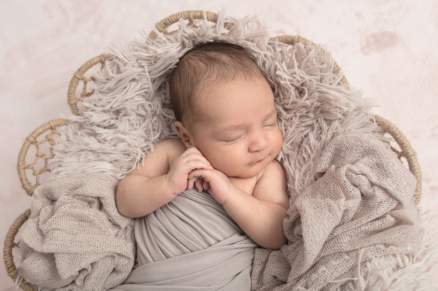 newborn wrapped in taupe and placed on an open knit blanket on top of a taupe colored flokati in a basket, baby clasping hands together