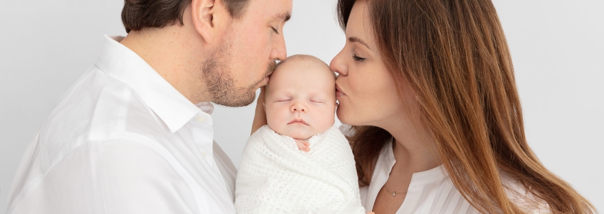 Studio posed newborn baby portrait session all in white, baby is cradled in-between mom and dad, kissing him on the cheeks