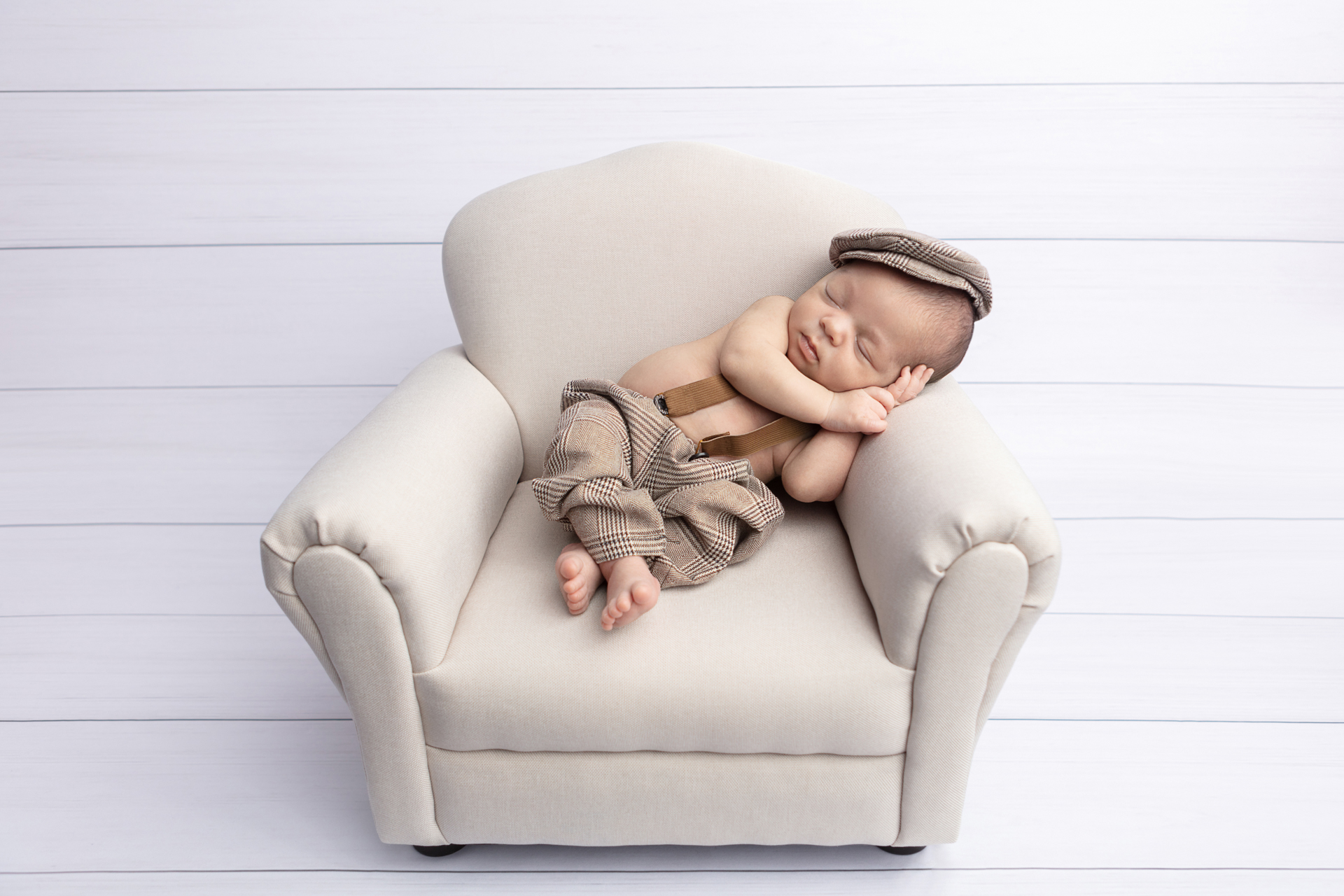 baby boy propped on a neutral oversized chair, wearing a cap and suspenders; CT newborn photography
